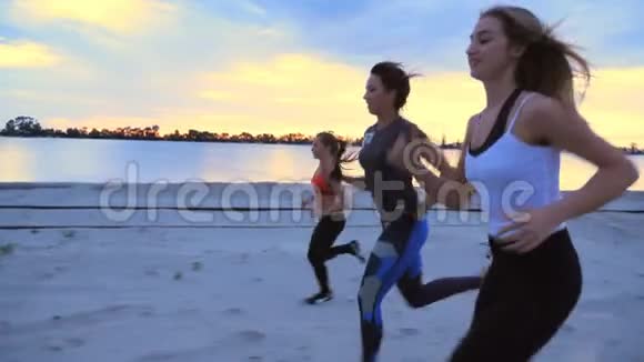 清晨的黄昏时分三个穿着运动服的女运动员沿着货运港的沙墩慢跑视频的预览图