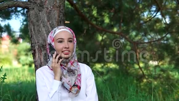一位现代穆斯林妇女在智能手机上讲话她坐在公园的一棵树旁视频的预览图