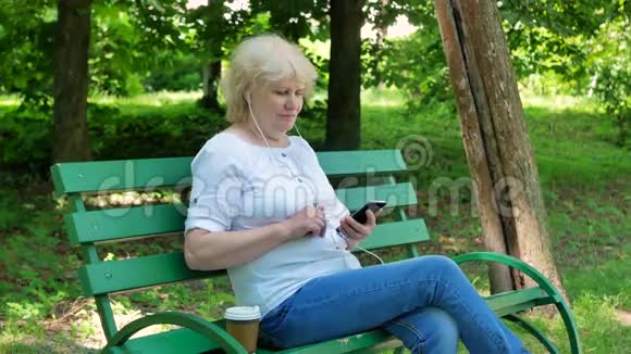 快乐的老妇人坐在公园的长凳上用智能手机带耳机喝咖啡高级女性视频的预览图