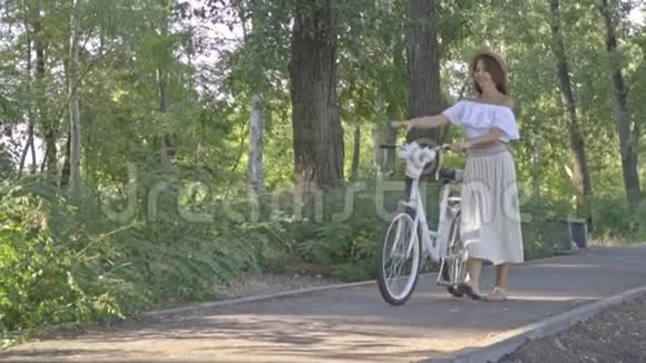 一个穿着白色裙子和带草帽的衬衫的浅黑色女孩正在公园白色城市自行车的道路上滚动视频的预览图