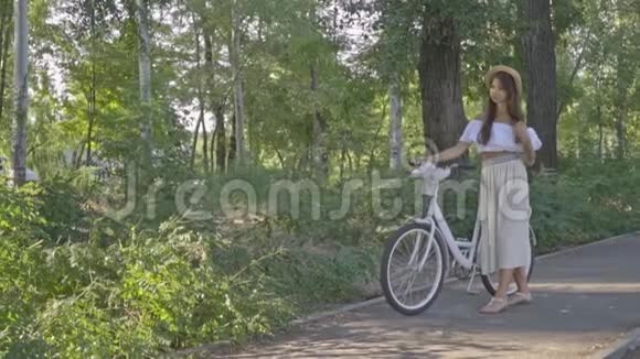 一个穿着白色连衣裙戴着纹身的帽子的浅黑肤色的美女调整了头发手里拿着一辆城市自行车慢动作射击视频的预览图