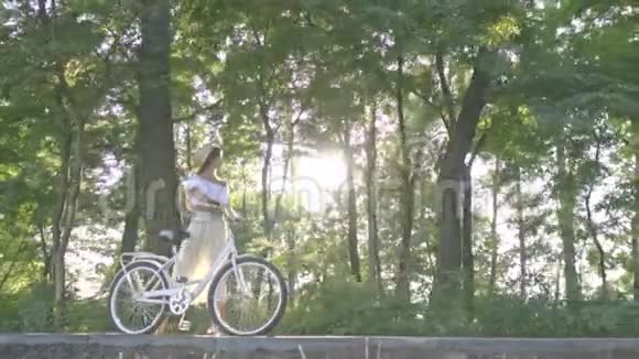 一个穿着白色裙子一件衬衫一辆白色城市自行车在阳光下翻滚的甜美黑发女人在公园的小路上视频的预览图