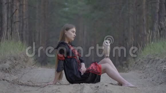 一个穿着黑色连衣裙的长发女孩光着脚坐在一条奉承的小路上膝盖上坐着一只浅棕色的猫头鹰慢吞吞的视频的预览图