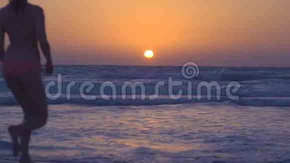 快乐的女孩正在海边奔向日落穿着粉红色比基尼的年轻女子在平静的海洋中奔跑跳跃视频的预览图