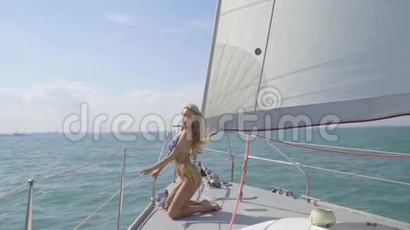 一个身穿蓝色比基尼的苗条金发女郎跪在游艇的船头上在阳光下驶向城市视频的预览图