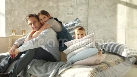 一个有两个孩子的年轻父亲坐在床上一个大家庭的早晨女孩拥抱父亲男孩玩视频的预览图