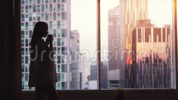 日落时年轻漂亮的女人在窗边喝着咖啡欣赏着令人惊叹的城市风景慢动作3840x2160视频的预览图