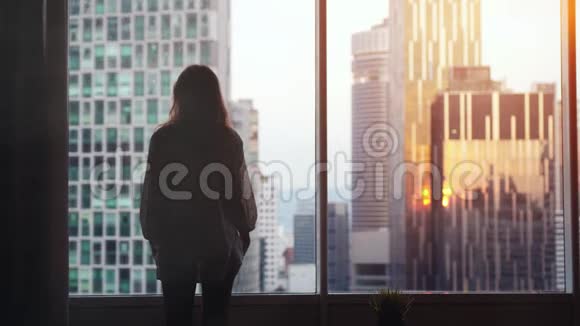 夕阳西下年轻忧郁的女人站在大窗户旁望着她在城市建筑上的公寓慢动作视频的预览图