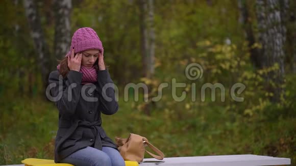 秋天公园里坐在长凳上头痛的不开心的漂亮女人慢动作视频的预览图