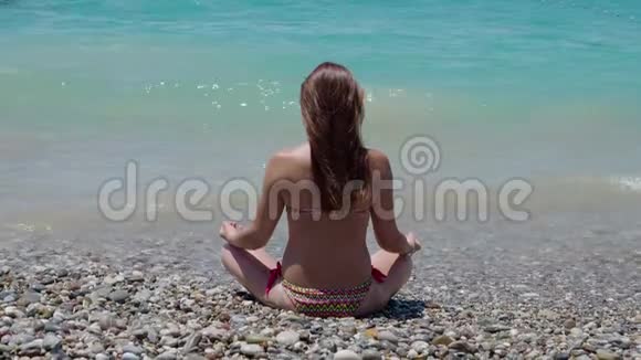 美丽的年轻女孩坐在海滩上的莲花姿势与她的背部4K视频的预览图