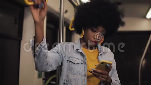 漂亮的非裔美国年轻女性握着扶手在公共交通中使用智能手机晚上视频的预览图
