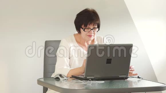 老妇人坐在桌旁手提电脑通过信使应用程序Skype使用计算机交谈微笑着挥手视频的预览图