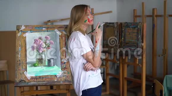 一位年轻迷人的艺术家手里拿着一把刷子脸上画着一张脸站在画架上视频的预览图