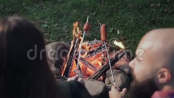 在篝火上烤香肠露营者烤香肠游客们正坐在火焰旁边视频的预览图