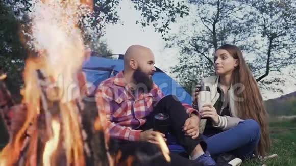 幸福的年轻夫妇坐在帐篷镇的炉火旁旅行者在森林里休息喝一杯热茶视频的预览图