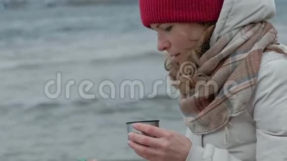 年轻女子穿着暖和的衣服坐在海边坐在木盘管上用电话从热水瓶里喝热茶视频的预览图