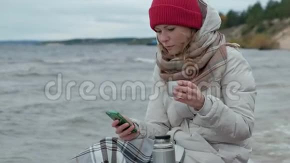 年轻女子穿着暖和的衣服坐在海边坐在木盘管上用电话从热水瓶里喝热茶视频的预览图