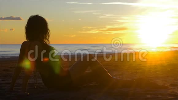 在日落时分年轻的黑发女人轻松地坐在沙滩上视频的预览图