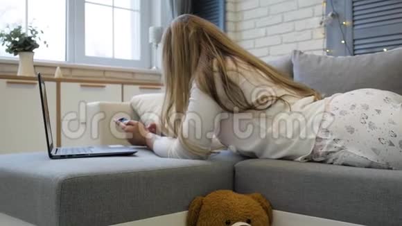 有智能手机和笔记本电脑的年轻女子躺在家里的沙发上视频的预览图