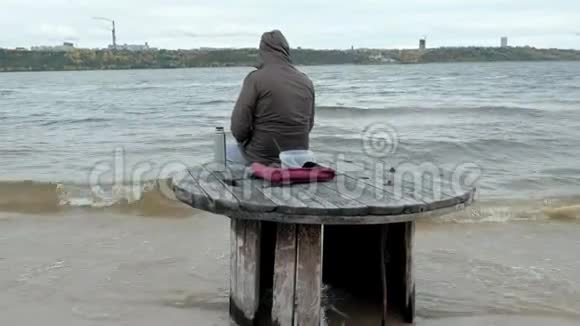 穿暖和衣服的年轻人坐在海边坐在木盘管上用热水瓶喝热茶视频的预览图