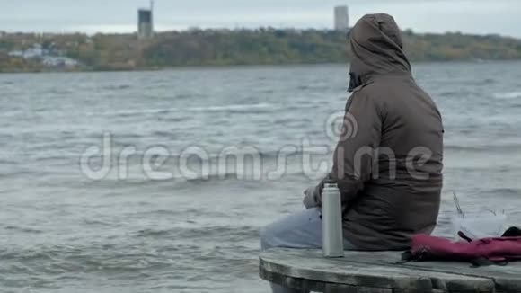 穿暖和衣服的年轻人坐在海边坐在木盘管上用热水瓶喝热茶视频的预览图