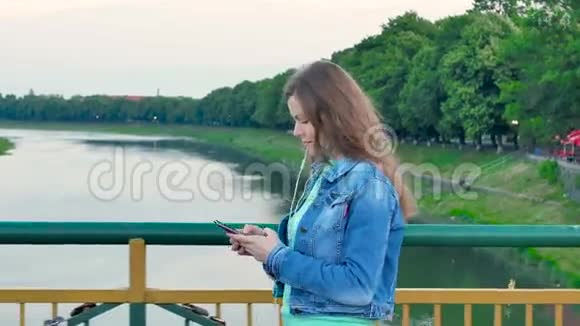穿着一件棉夹克的漂亮女孩穿过河上的桥用智能手机带耳机视频的预览图