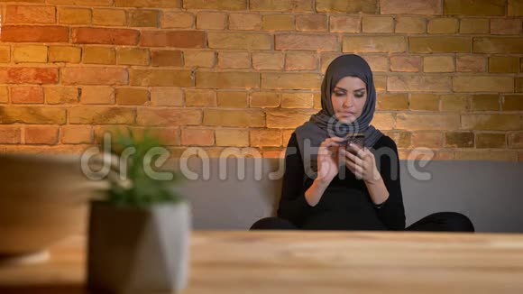 在室内沙发上坐着的穆斯林成年女性戴着头巾在手机上发短信视频的预览图