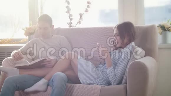 快乐的年轻情侣坐在沙发上男生看书女生用电话视频的预览图