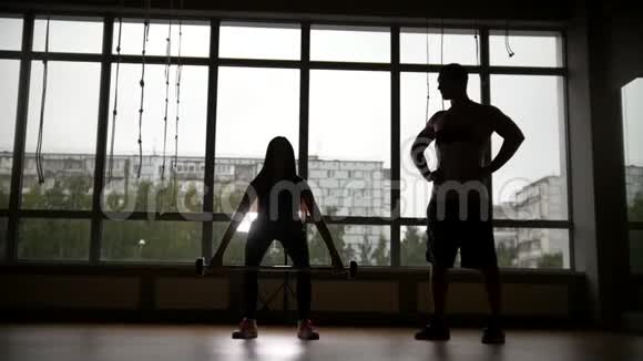 女孩和教练在健身房举起杠铃剪影慢动作滑块窗外多云的雨天视频的预览图