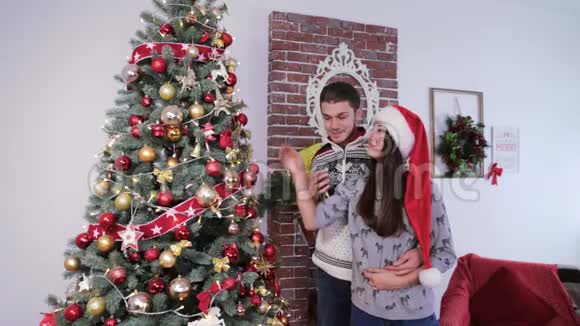 可爱的年轻夫妇用毛衣和圣诞老人红帽子装饰圣诞树视频的预览图
