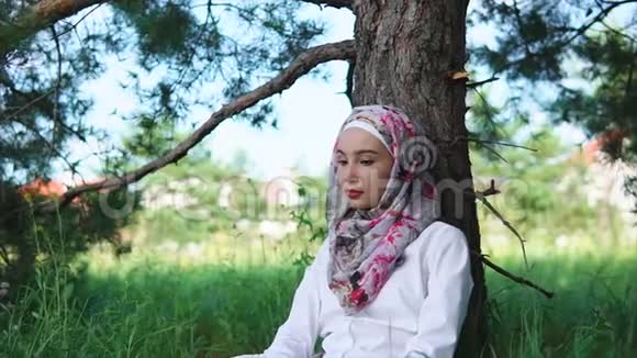 一个戴着穆斯林头巾的女人坐在公园树旁的草地上视频的预览图
