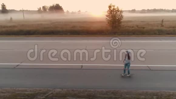 空中飞行一个年轻的女孩在黎明时骑在滑板上沿着一条荒芜的高速公路行驶视频的预览图