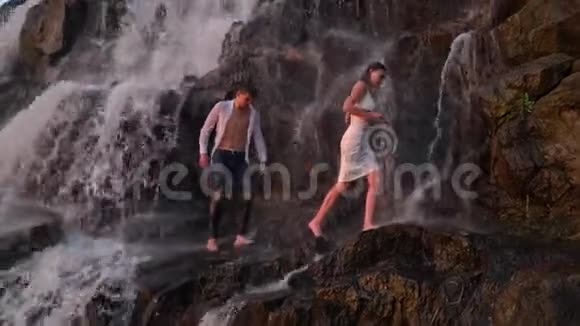 日落时分一个穿着白色衣服的男人和一个女孩整齐地在瀑布下的岩石中穿行视频的预览图