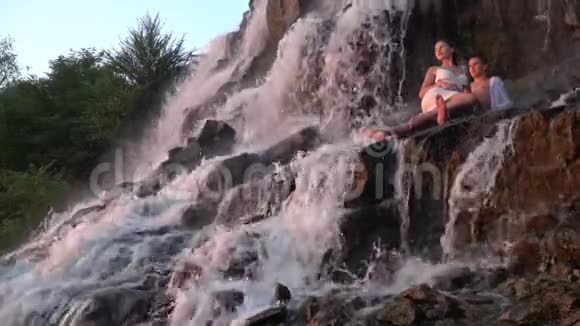 浪漫的日落为一对坐在瀑布下岩石上的年轻人慢动作视频的预览图