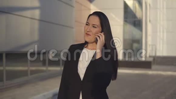 阳光明媚的一天穿着黑色西装的年轻女商人在办公室中心的背景下在智能手机上交谈视频的预览图