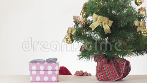 戴着圣诞老人帽子的美丽顽皮的少女在白色背景的新年树下偷偷地打开礼物视频的预览图