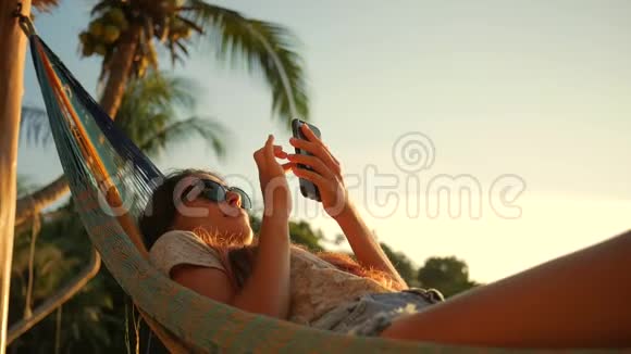 放松的混合种族年轻女子看手机吊床附近的海边日落戈芬加视频的预览图