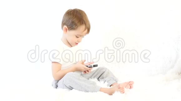 快乐的孩子在白色背景下玩智能手机一个孩子坐在白色沙发上视频的预览图