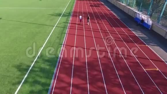 在阳光明媚的日子里穿着运动服的白人和非裔美国人在跑道上奔跑在空中拍摄视频的预览图