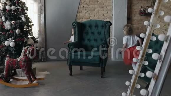 两个穿着红色裙子的女孩围着一张绿色的椅子跑来跑去椅子停在靠近圣诞节的客厅里视频的预览图