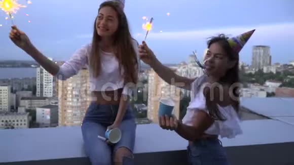 两个开朗的黑发女人吹着明亮的帽子吹着口哨庆祝生日在大楼的屋顶上靠在屋顶上视频的预览图