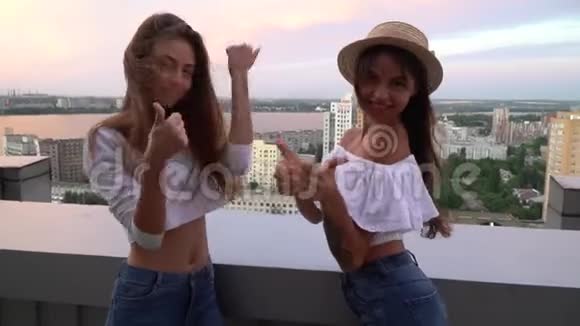 两个长着长发的活泼的黑发女孩其中一个戴着草帽两人都竖起大拇指站在屋顶上视频的预览图