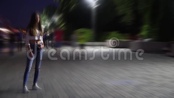 晚上一个穿着衬衫和牛仔裤的长发孤女站在广场上散步的人们中间视频的预览图