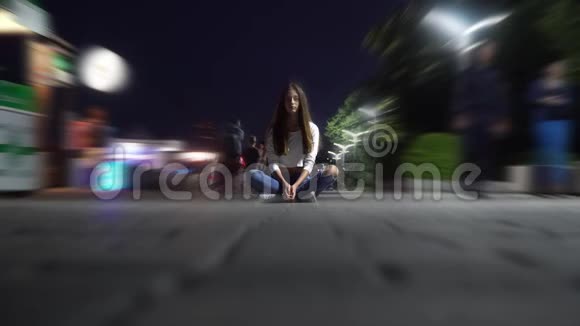 晚上一个穿着衬衫和牛仔裤的女孩坐在广场中央人们在那里散步和微笑镜头模糊视频的预览图