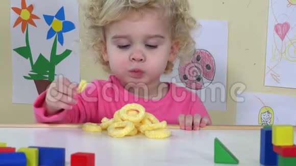 可爱的脸蛋女孩吃玉米圈坐在她的桌子旁边玩具和图纸视频的预览图