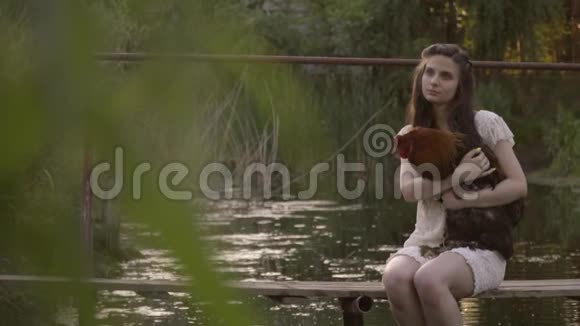 美丽的农村女孩穿着白色蕾丝连衣裙坐在河边的木桥上抱着公鸡平静的农村生活在大自然中视频的预览图