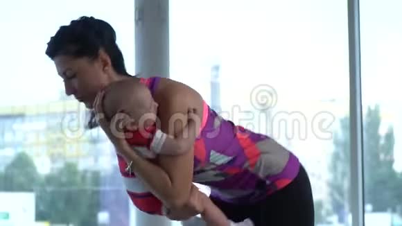 妈妈抱着孩子摇着他的背弯着腰不停地站在健身房做瑜伽视频的预览图