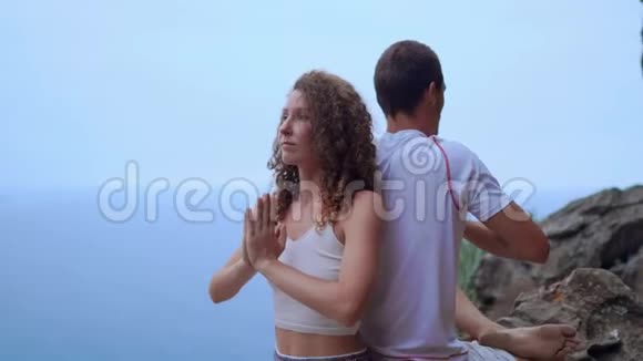 男人和女人坐在山顶的岩石上背靠背冥想在大海的背景下做瑜伽视频的预览图
