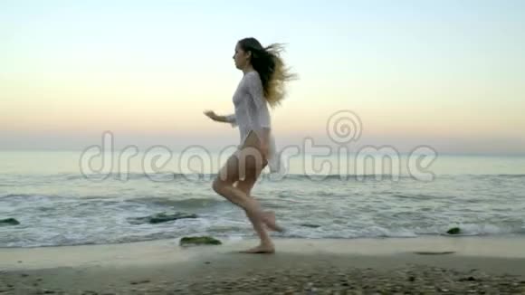穿着白色短裙的年轻女孩在夕阳的背景下沿着海边奔跑慢镜头拍摄视频的预览图