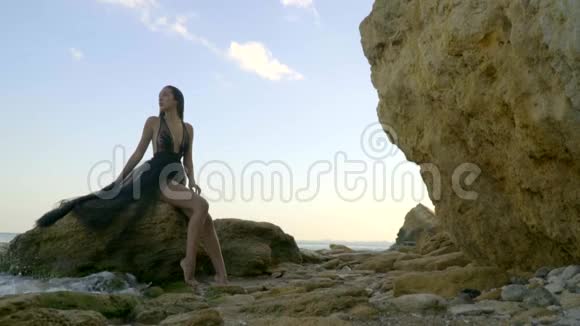 一个忧郁的黑发女人穿着一件通风的黑色连衣裙坐在一块破碎的岩石上海浪在盐滩上拍打着视频的预览图